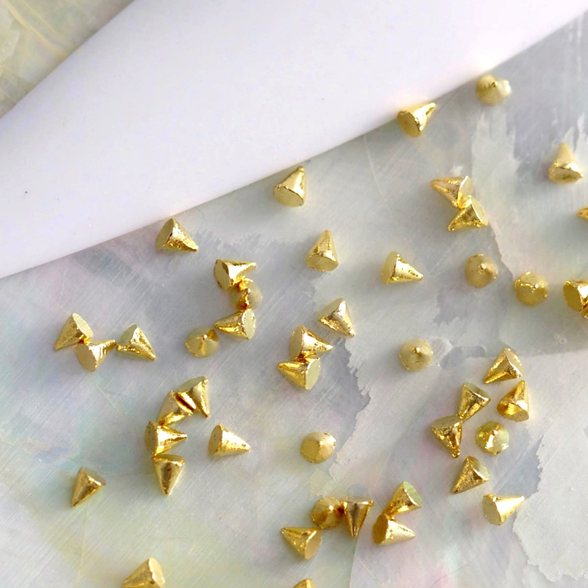 Warm gold mini cone spike studs and matching round metal accents spread across a light, textured surface next to a white nail tip, illustrating the metallic hardware style and how they can be combined in nail art.