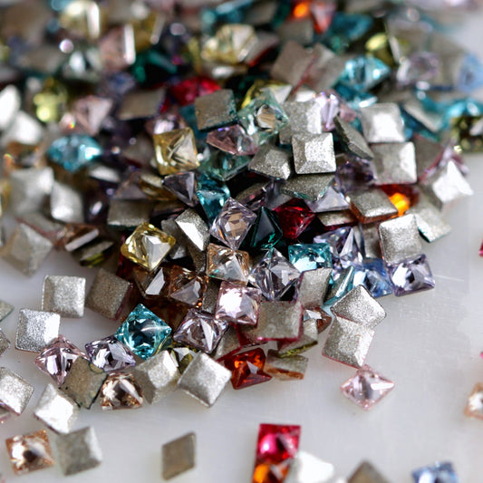 A macro view of 1.5mm square zircon nail-art stones scattered together, with faceted fronts in assorted colours and flat adhesive backs visible. Each package contains a random colour mix, so the colour balance may differ.