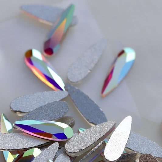 Close-up of multiple teardrop-shaped rhinestones with iridescent, color-shifting surfaces and some glittery silver rhinestones scattered on a light background.