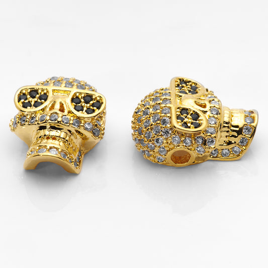 Large gold skull beads paved with crystal and jet CZ stones shown on a white nail tip against a white background.