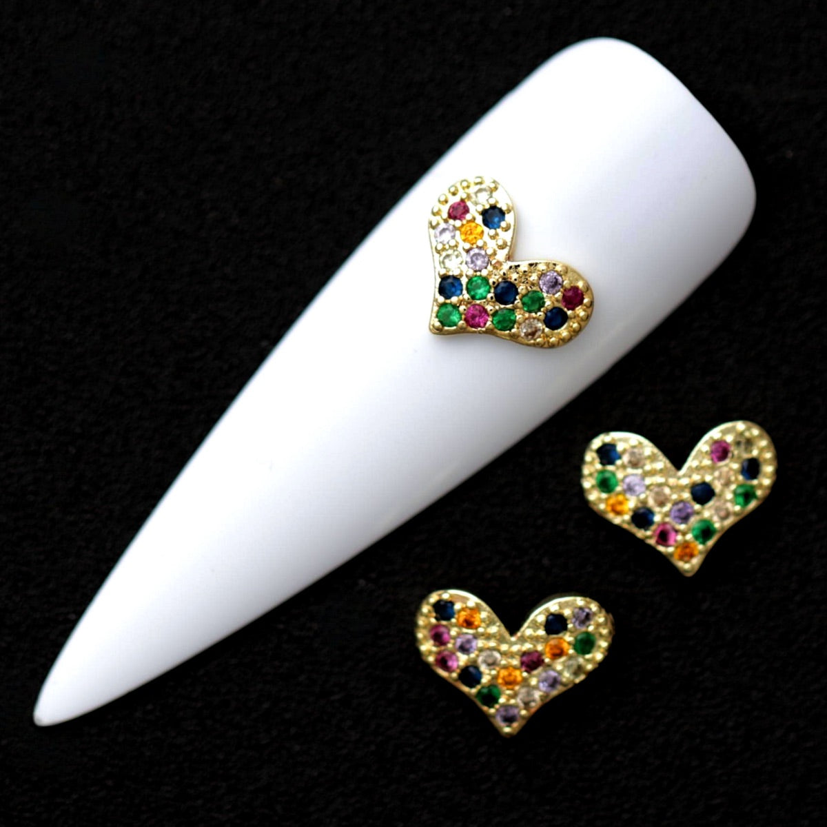 Gold 10×8 mm puffed heart nail charms fully paved with multicolor CZ stones displayed on a white nail tip against a black background