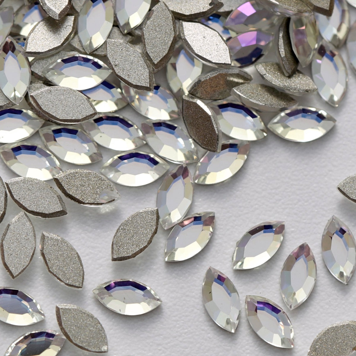 A detailed arrangement of K9 Crystal Flatback Navette – Crystal Moonlight rhinestones displayed on a white background. The slim, leaf-like crystals reflect cool crystal light with subtle blue, lavender, and silver highlights, while the silver backing enhances their brilliance. Ideal for geometric nail designs, bridal embellishments, jewelry, fashion accessories, and refined DIY projects.