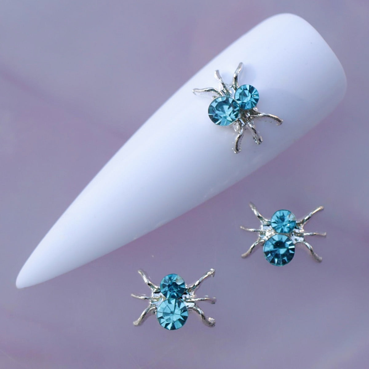 Silver-tone spider nail charm featuring delicate curved legs and two faceted aquamarine crystal rhinestones. The blue crystals reflect aqua, turquoise and light-blue tones, making this distinctive spider decoration ideal for Halloween manicures, ocean-inspired designs, mermaid nail art, gothic looks and creative accent nails.