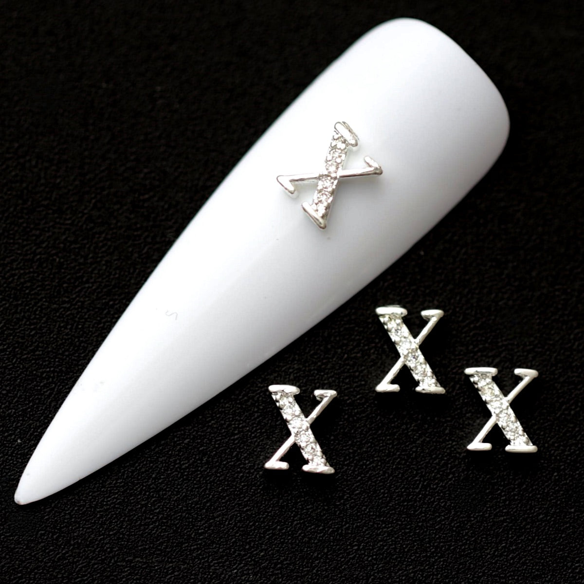 Silver X letter nail charms paved with crystal cubic zirconia on a white nail tip against a black background