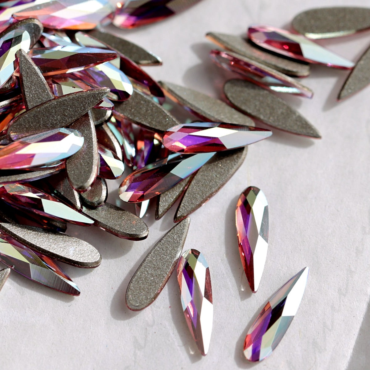 A pile of faceted teardrop-shaped Swarovski 2304 Light Rose AB flatback rhinestones with a metallic rose gold edge and an iridescent, color-shifting surface that reflects pink, purple, and blue tones, displayed on a light gray textured background.