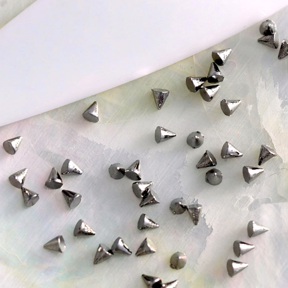 Small metallic gunmetal spike studs scattered on a pale iridescent surface with a white curved shape in the upper left.