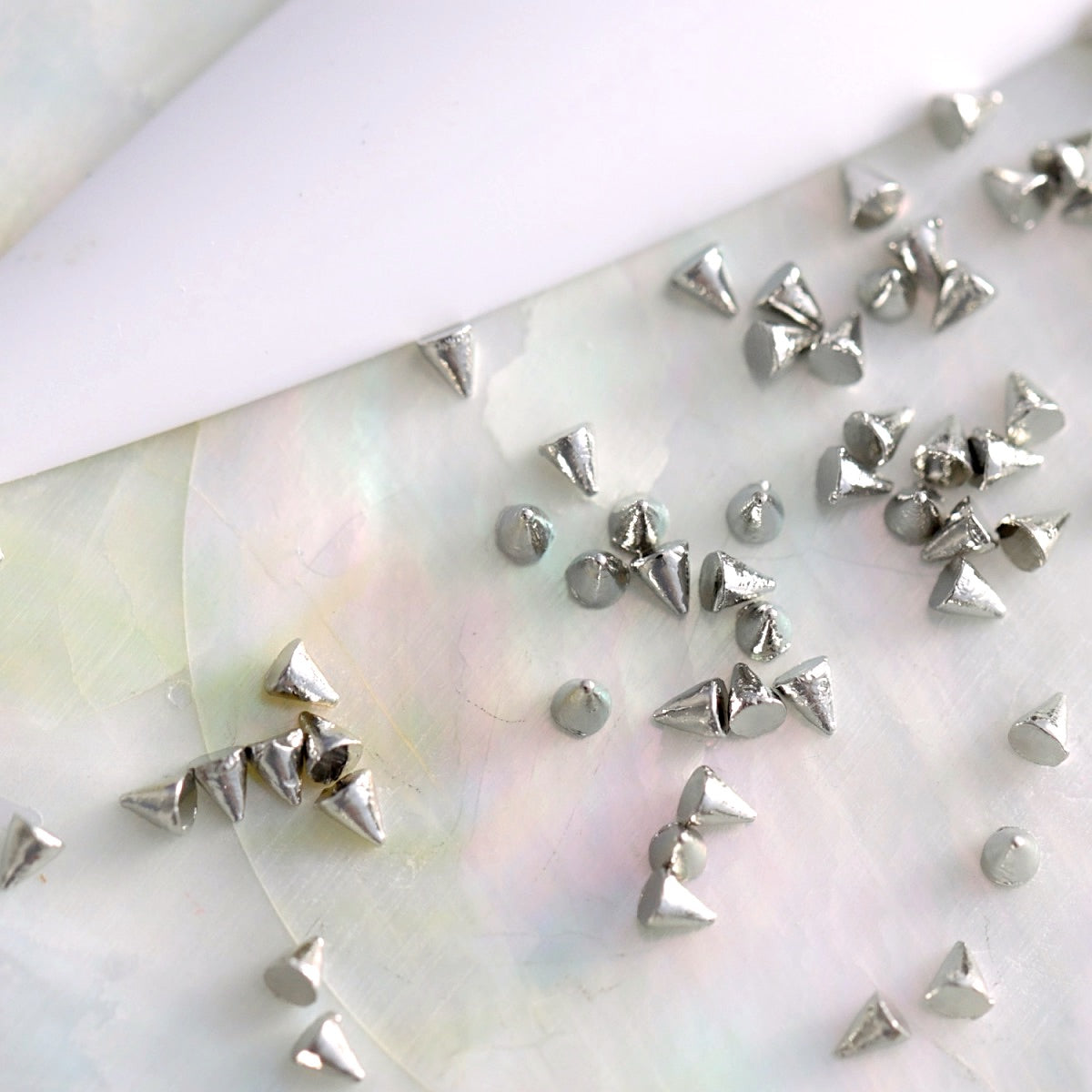 Close-up of silver mini cone spike studs arranged on a soft, iridescent surface beside a white nail tip, showing their 3D shape and flat backs for nail art application.