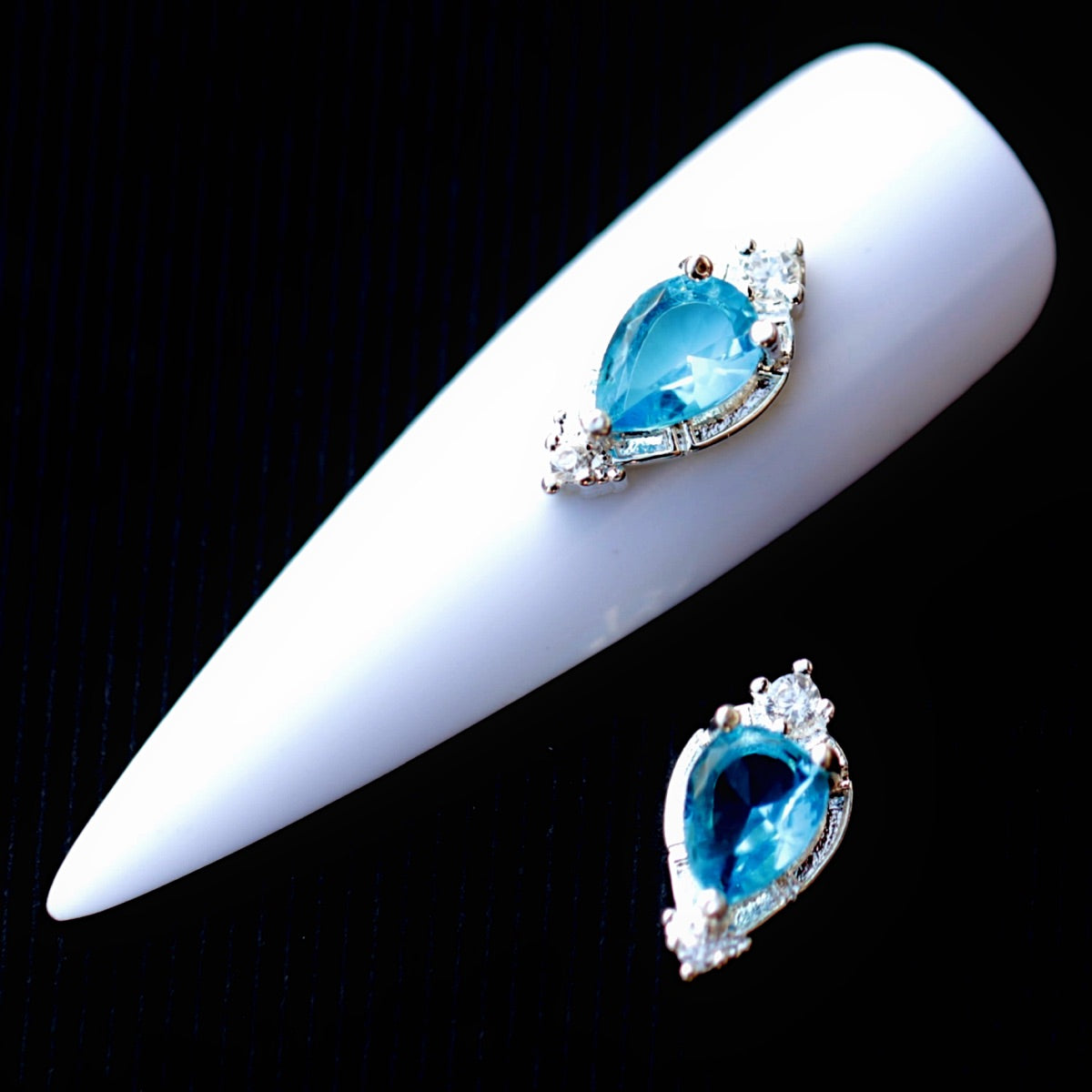 Decorative nail charm featuring a large faceted aquamarine teardrop-shaped cubic zirconia crystal in the centre, surrounded by clear CZ accents and a silver-tone setting. The jewellery-inspired nail decoration is suitable for bridal manicures, ocean-themed designs, mermaid nail art, summer looks and elegant statement accent nails.