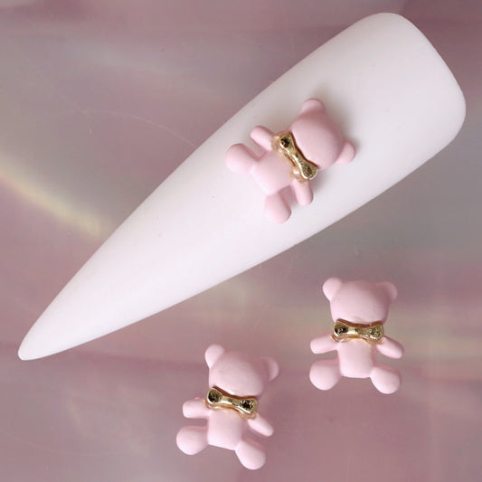 Baby pink teddy bear nail charm featuring rounded ears, small limbs and a shiny gold bow around its neck. This cute pastel decoration is ideal for kawaii manicures, soft-girl nail art, romantic designs, baby-themed looks and playful accent nails.