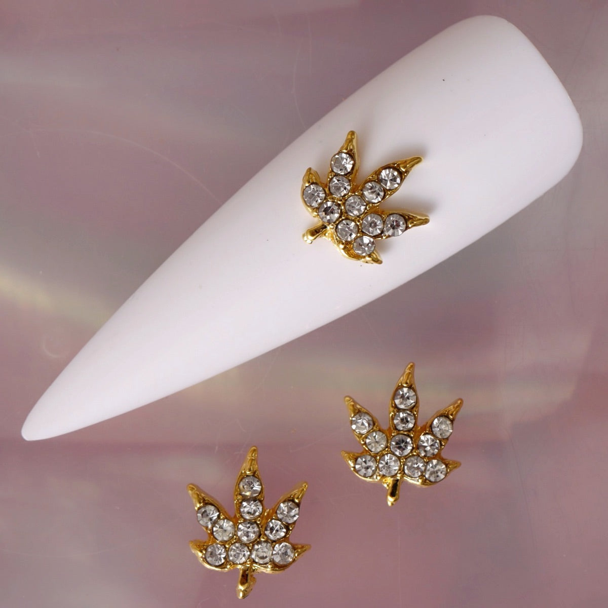 Cannabis leaf-shaped nail charm with a warm gold-tone base and clear transparent rhinestones arranged across the pointed leaf sections. This distinctive botanical nail decoration is ideal for festival manicures, themed nail art, statement accent nails and creative designs.
