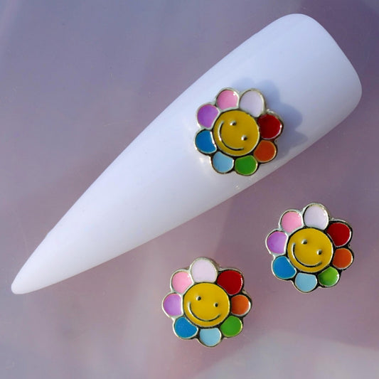 Colourful flower nail charm featuring a bright yellow smiley face in the centre and rounded petals in pink, purple, white, red, orange, green, blue and light blue. This cheerful novelty nail decoration is ideal for kawaii manicures, colourful summer nail art, festival looks, birthday nails and playful accent designs.