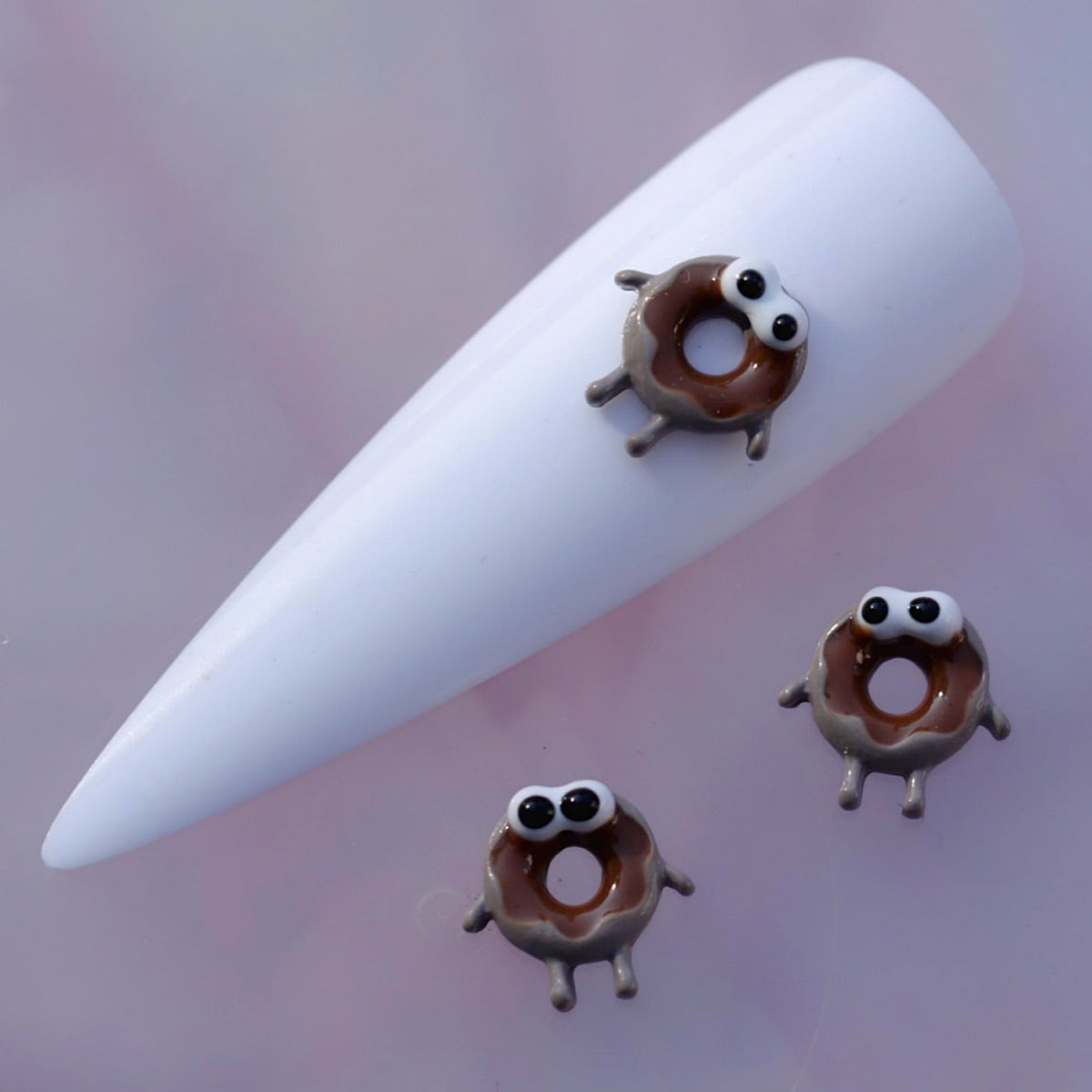 Three-dimensional donut nail charm with glossy chocolate-brown glaze, a lighter baked base, a central hole, funny eyes and tiny decorative legs. This playful dessert-themed decoration is ideal for kawaii manicures, bakery-inspired nail art, food designs and fun accent nails.