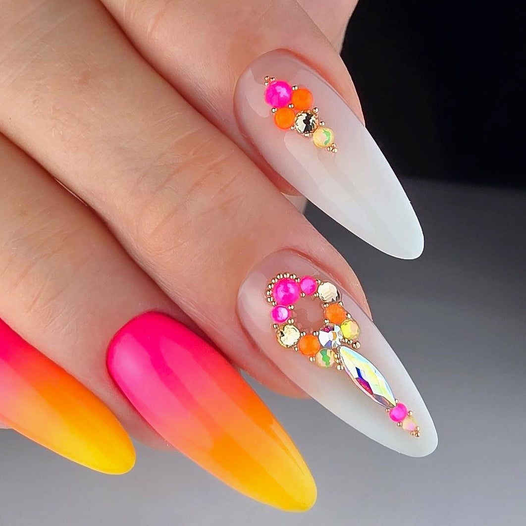 Close-up of a hand with long stiletto nails decorated with colorful nail art. One nail has a bright pink-to-yellow gradient, another has a translucent white base with orange and gold bead accents, and a third has a matching white base with a rhinestone circle and iridescent jewel.