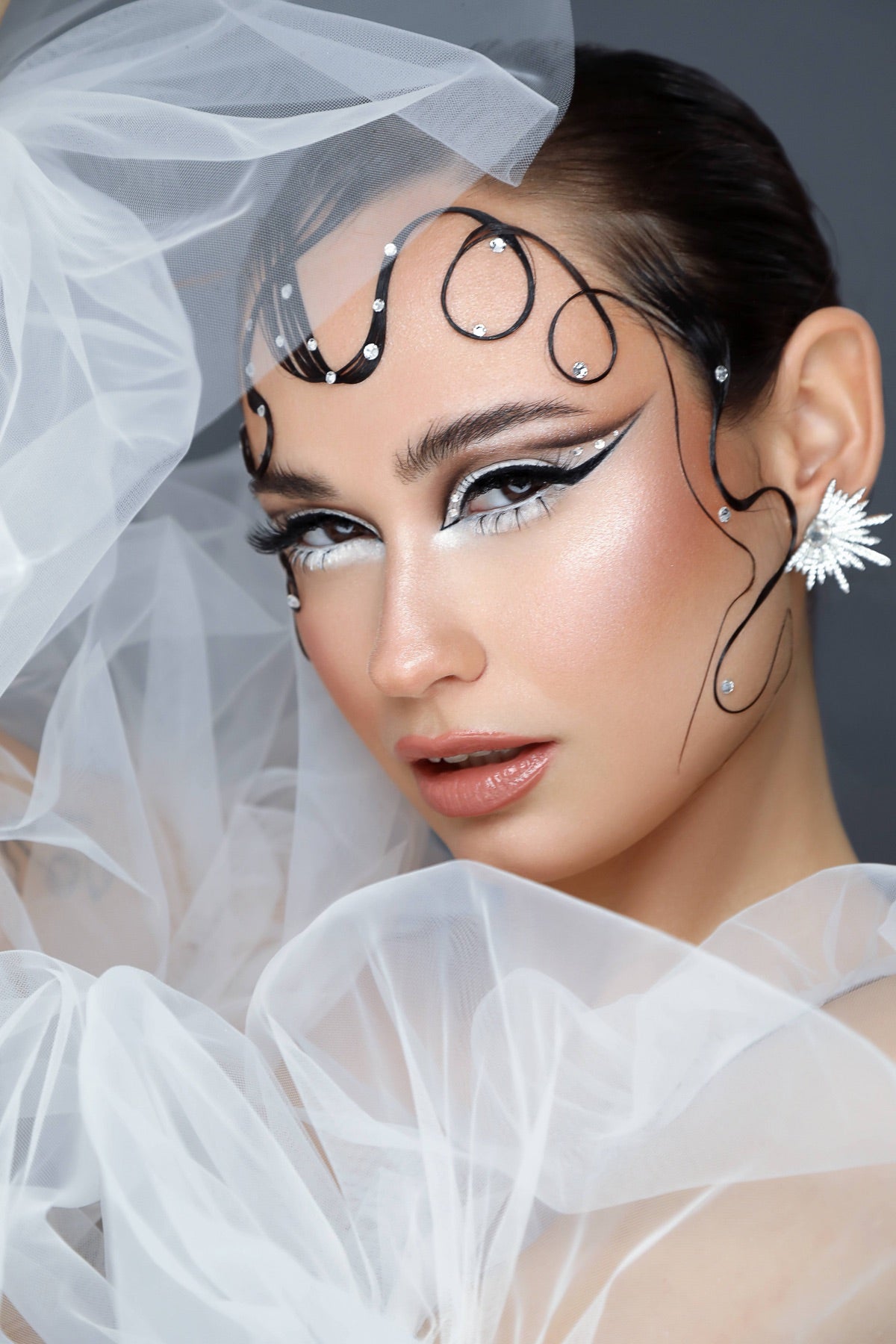 A close-up portrait of a woman with dramatic black and white eye makeup, decorative black wire headpiece with small rhinestones, a white flower earring, and sheer white tulle fabric around her head and shoulders against a dark gray background.