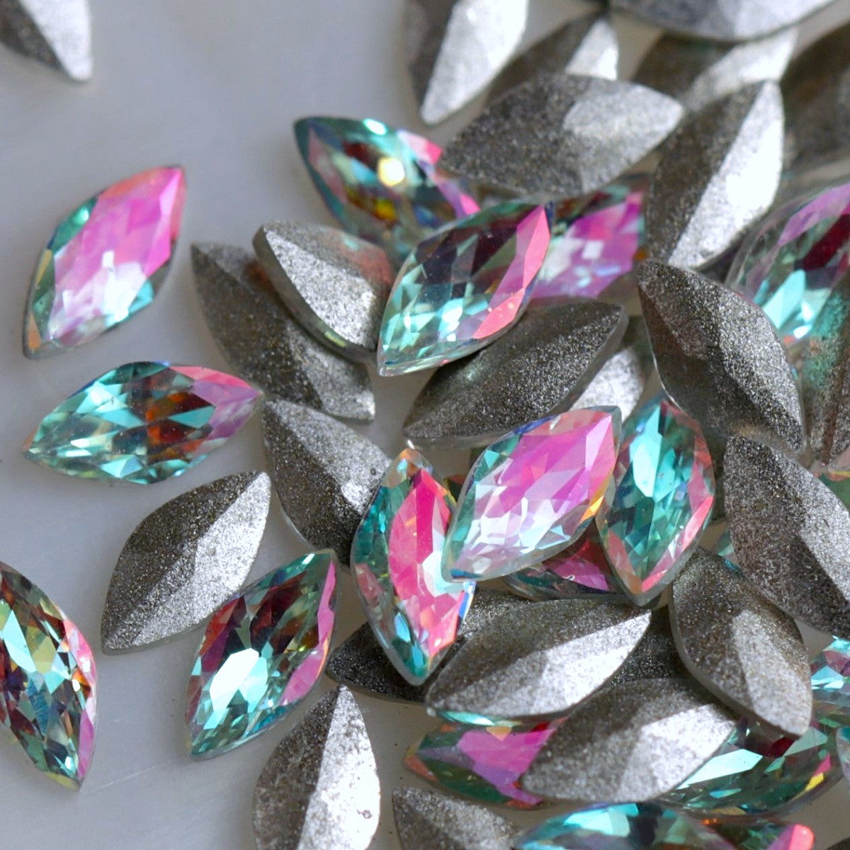 Faceted glass navette stones with a pale aqua-turquoise centre and luminous pink iridescent shimmer. The stones reflect mint, blue, lilac, silver and soft rainbow tones depending on the light. Their backs are slightly convex rather than completely flat, making them suitable for experienced nail artists creating floral, bridal, mermaid and detailed crystal nail-art compositions.