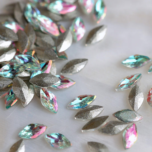 Close-up of loose marquise-shaped iridescent rhinestones scattered on a light surface, showing holographic rainbow reflections.