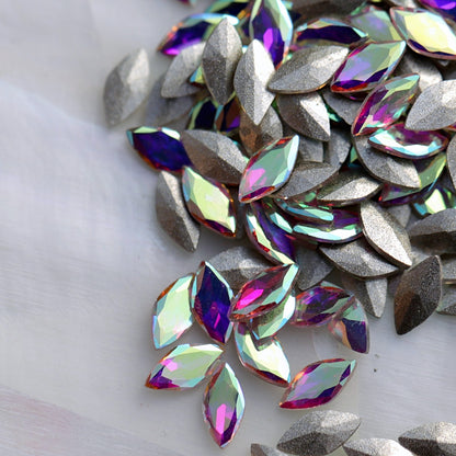 Faceted glass navette stones in Crystal AB with a clear glass base and multicolour Aurora Borealis reflections. The pointed oval stones have a slightly convex underside rather than a fully flat back, making them suitable for experienced nail artists using strong gel adhesive in floral, bridal, mermaid and detailed nail-art compositions.