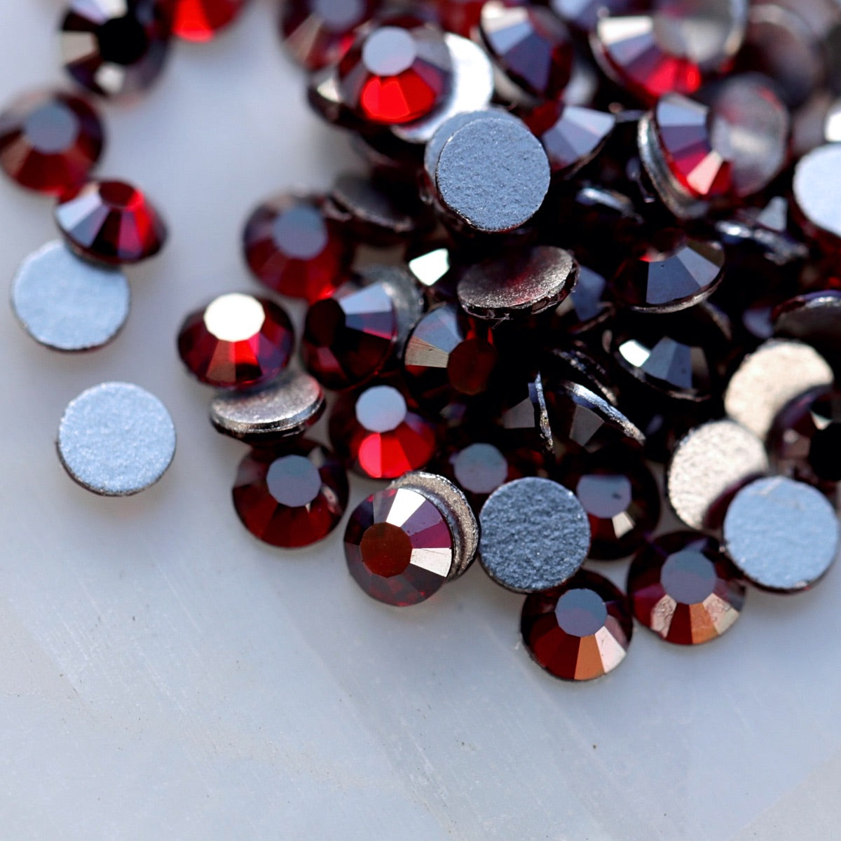 Faceted glass flatback rhinestones in a deep Dark Siam burgundy-red shade with a special metallic silver reflective shimmer visible from selected angles. The stones show rich dark ruby-red colour with cool silver, steel-grey and graphite-like flashes, making them ideal for gothic, Christmas, winter, party and dark romantic nail art.