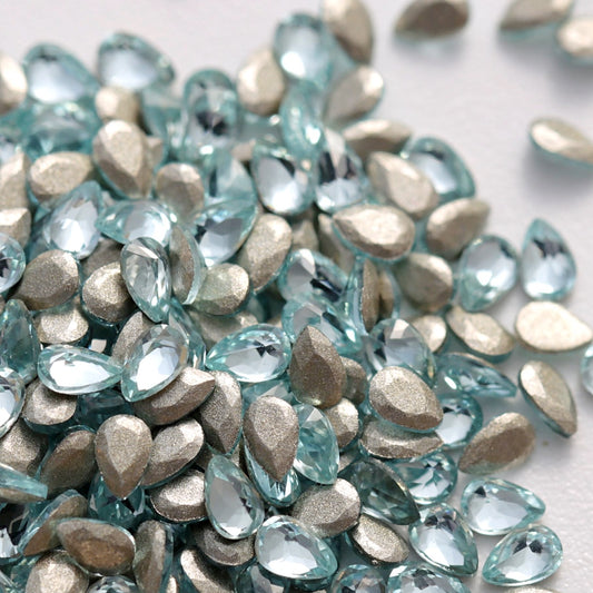 A detailed close-up of Aquamarine teardrop-shaped zircon crystals displayed together with their flatback foil surfaces visible. The refreshing blue tone, elegant drop shape, and faceted finish create a luminous, multidimensional effect, making these crystals perfect for nail art, jewelry, accessories, and creative craft projects.