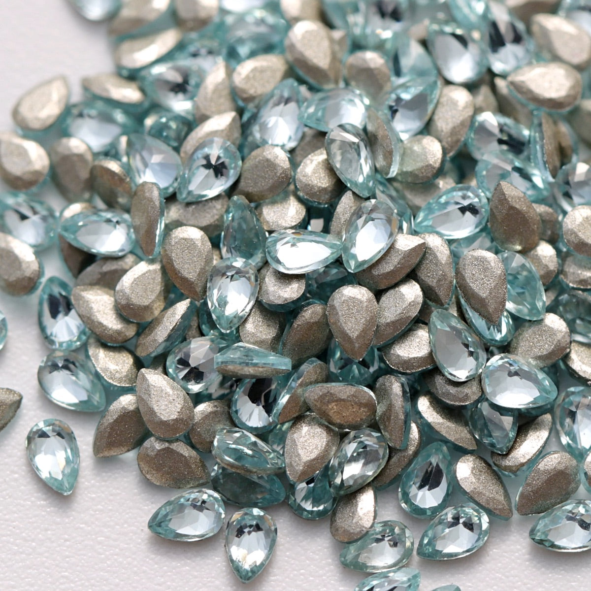 A close-up view of Aquamarine flatback zircon drop crystals arranged on a white surface. The delicate light-blue teardrop-shaped stones feature precision-cut facets and reflective foil backings that create a bright, elegant sparkle. Ideal for nail art, jewelry accents, fashion decoration, and creative DIY embellishments.