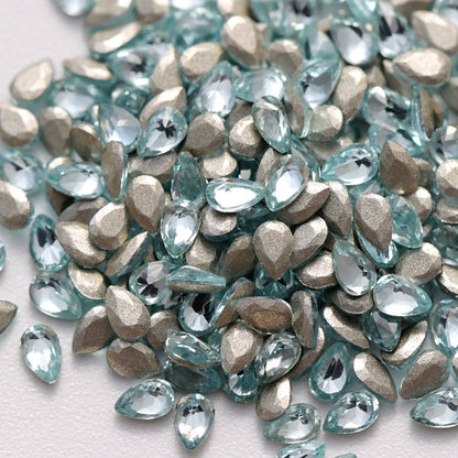 A close-up view of Aquamarine flatback zircon drop crystals arranged on a white surface. The delicate light-blue teardrop-shaped stones feature precision-cut facets and reflective foil backings that create a bright, elegant sparkle. Ideal for nail art, jewelry accents, fashion decoration, and creative DIY embellishments.