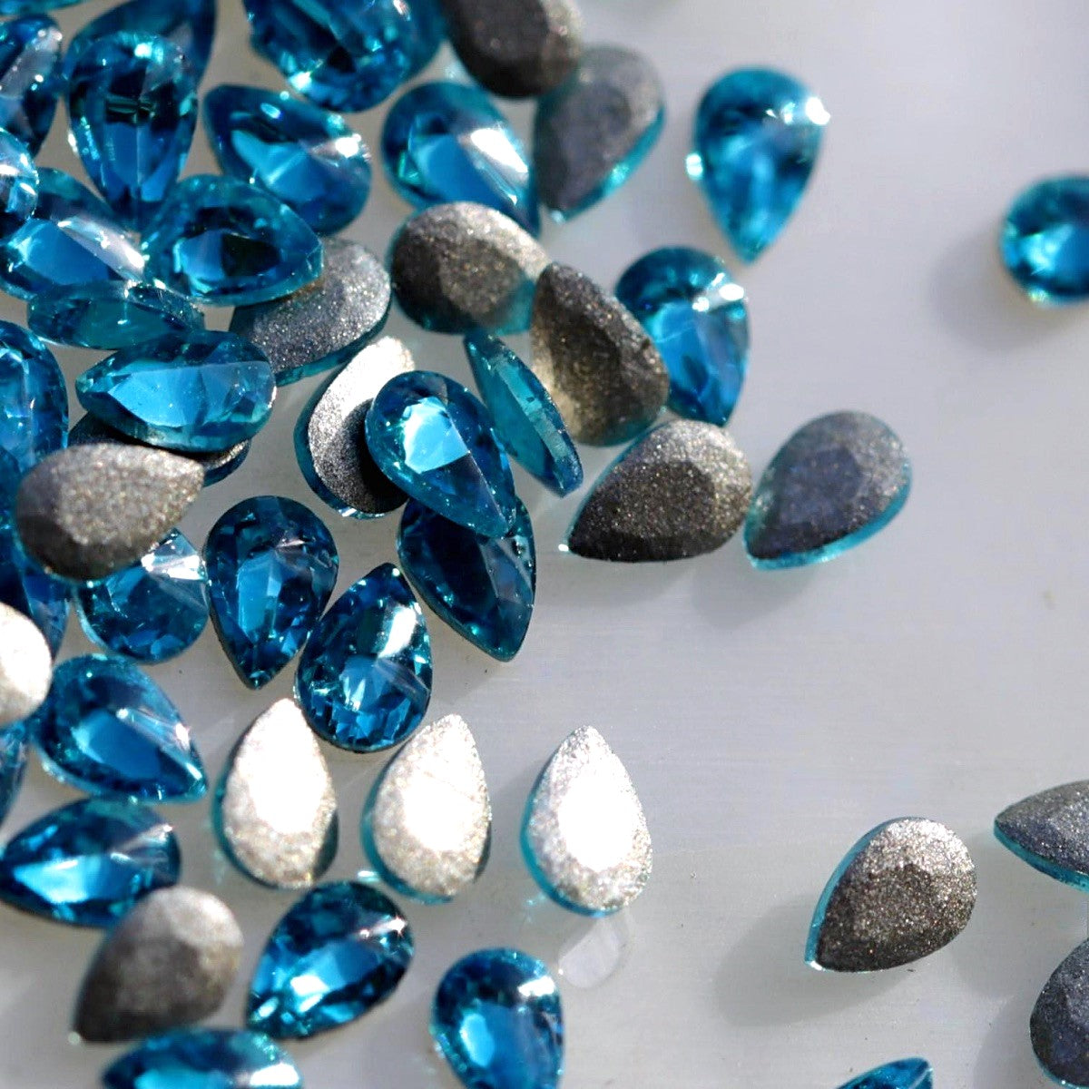 Flatback Zircon Nail Art Drop crystals in Caribbean Blue, measuring 3×2mm. The small teardrop-shaped crystals feature a precision-faceted front surface with bright turquoise, aqua, cyan, Caribbean blue, ocean-blue and icy reflections. Ideal for tropical, ocean-inspired, mermaid, floral, summer and creative nail art designs.