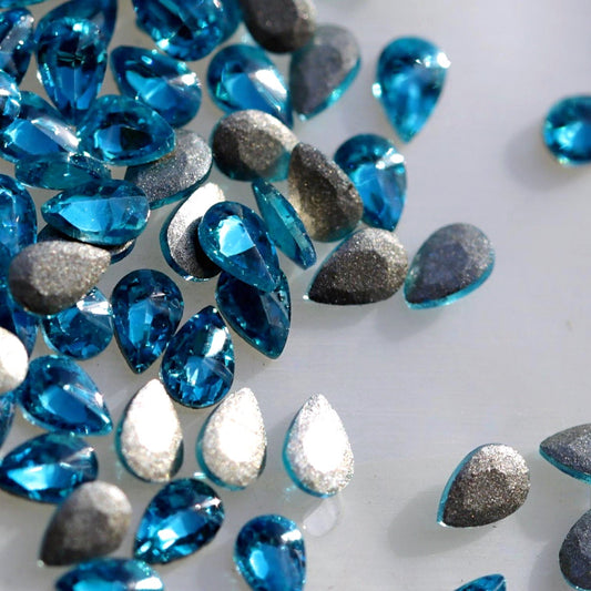 Flatback Zircon Nail Art Drop crystals in Caribbean Blue, measuring 3×2mm. The small teardrop-shaped crystals feature a precision-faceted front surface with bright turquoise, aqua, cyan, Caribbean blue, ocean-blue and icy reflections. Ideal for tropical, ocean-inspired, mermaid, floral, summer and creative nail art designs.