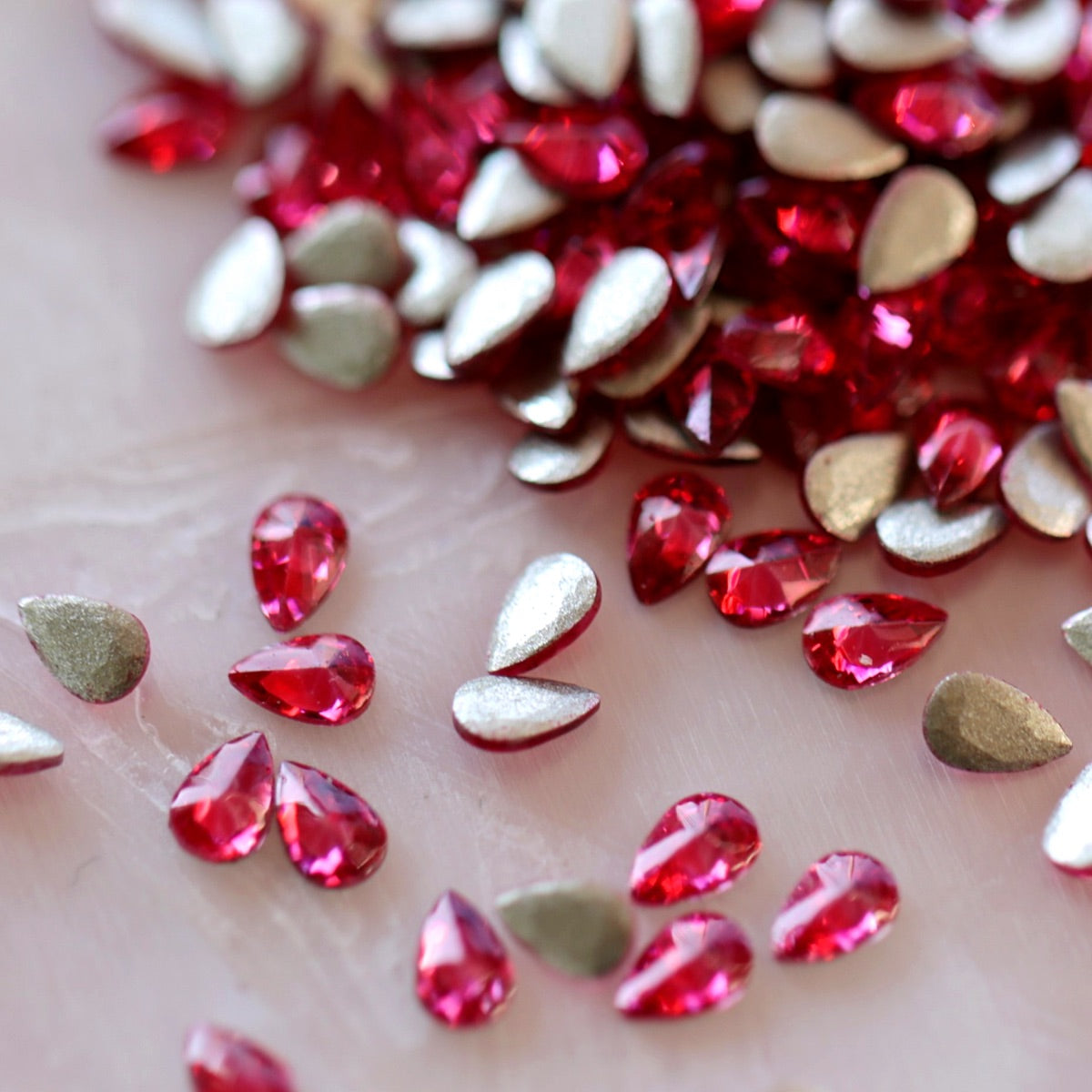 Flatback Zircon Nail Art Drop Magenta stones scattered on a light surface, displaying vivid pink teardrop crystals alongside their metallic foil backs.