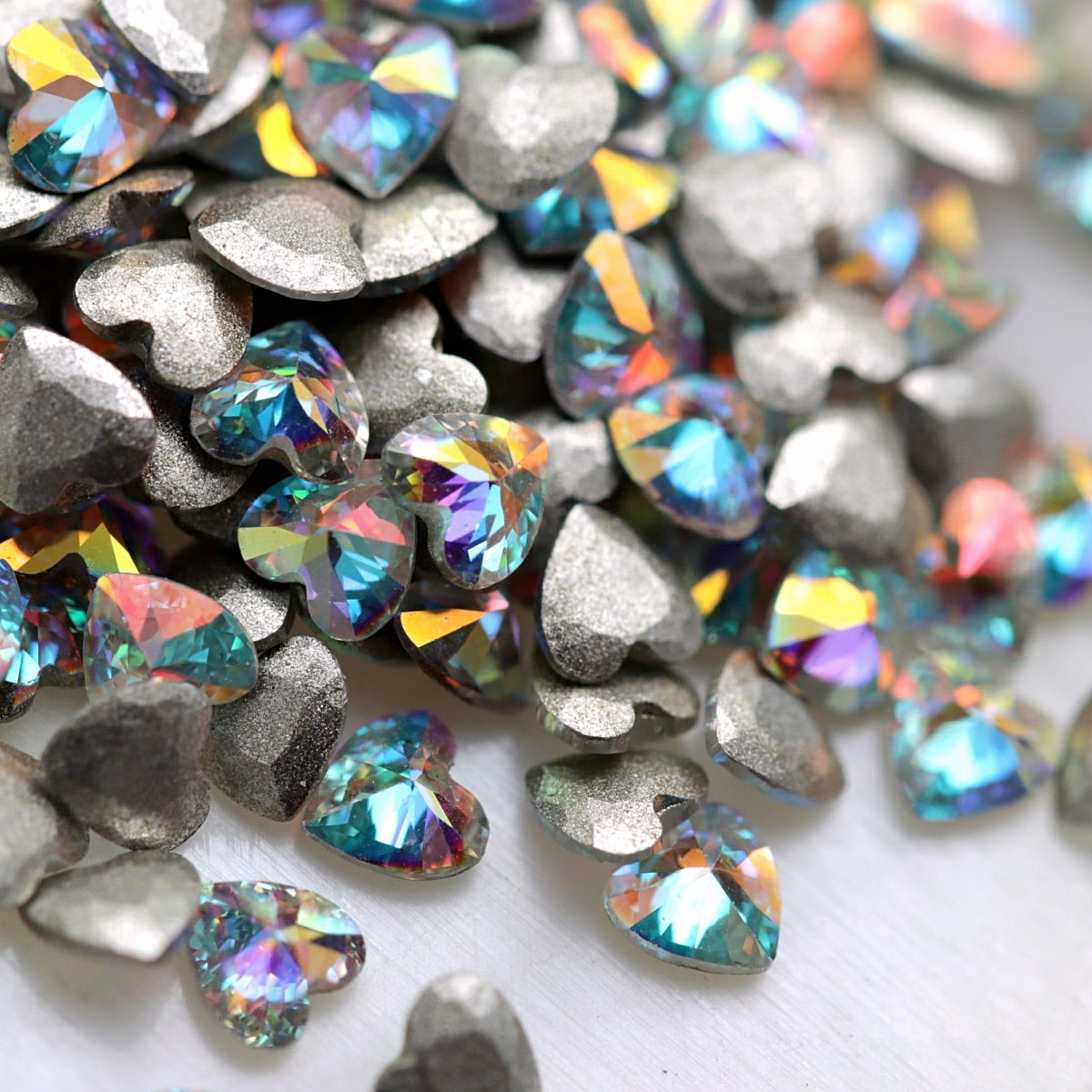 Close-up of 3×3mm Crystal AB faceted zircon heart decorations, showing clear crystal bases with aqua, pink, purple and rainbow Aurora Borealis reflections.