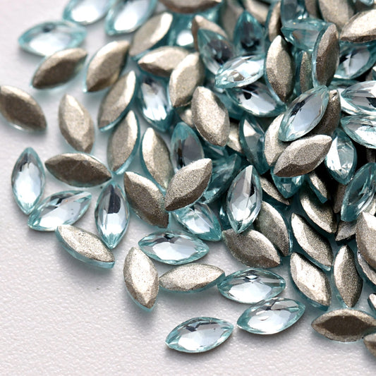 A close-up view of Aquamarine flatback zircon marquise crystals arranged on a white surface. The elongated navette-shaped stones feature a soft, refreshing light-blue color, precision-cut facets, and reflective flatback foil surfaces that create an elegant crystal sparkle. These versatile rhinestones are suitable for nail art, jewelry decoration, fashion accessories, and creative DIY embellishments.