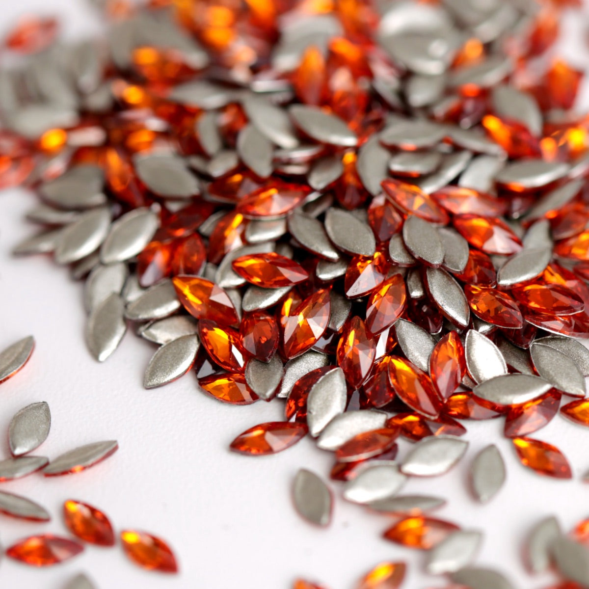 Flatback zircon nail art marquise crystals in vibrant Hyacinth red-orange. A pile of marquise-cut rhinestones in orange and silver, scattered on a white surface.