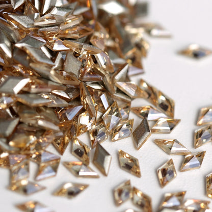 A close-up view of Light Peach flatback zircon rhombus crystals arranged on a white surface. The diamond-shaped stones feature a soft peach tone, precision-cut facets, and reflective foil backings that create elegant sparkle and depth. Ideal for geometric nail art, jewelry decoration, fashion accessories, and creative embellishments.