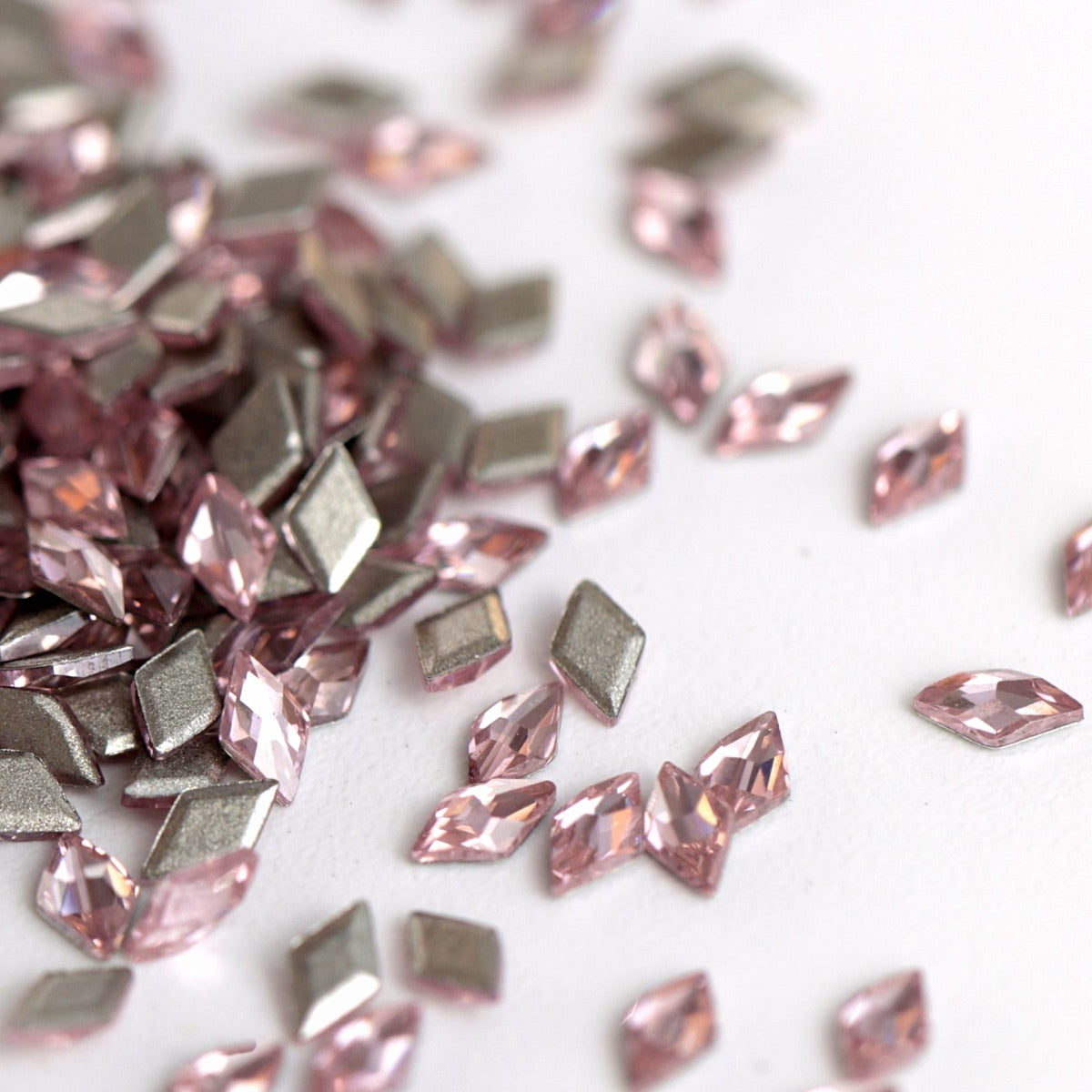 A close-up view of Light Rose flatback zircon rhombus crystals scattered across a clean white surface. The soft pink stones feature precise geometric cuts and reflective facets that create a delicate, elegant sparkle. Ideal for refined nail art, jewelry accents, fashion decoration, and creative DIY projects.