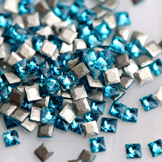 Close-up of 2mm Caribbean Blue faceted zircon square nail-art stones, showing their vivid blue-turquoise colour and flat adhesive backs.