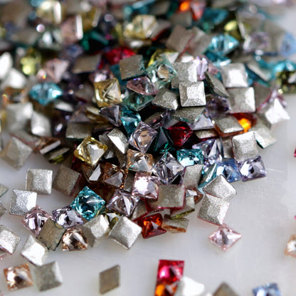 A macro view of 1.5mm square zircon nail-art stones scattered together, with faceted fronts in assorted colours and flat adhesive backs visible. Each package contains a random colour mix, so the colour balance may differ.