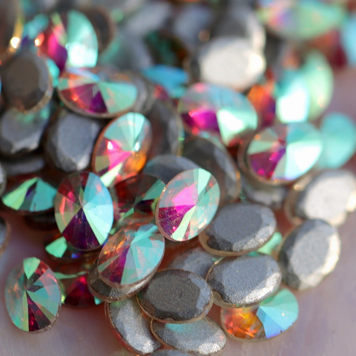 Piled oval shaped flatback zircons in AB coating showing rainbow refractions from green to pink on soft background for nail art embellishments