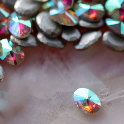 Scattered oval AB flatback zircon crystals with vibrant pink, teal, orange, and purple iridescent effects on pearlescent surface for nail decoration