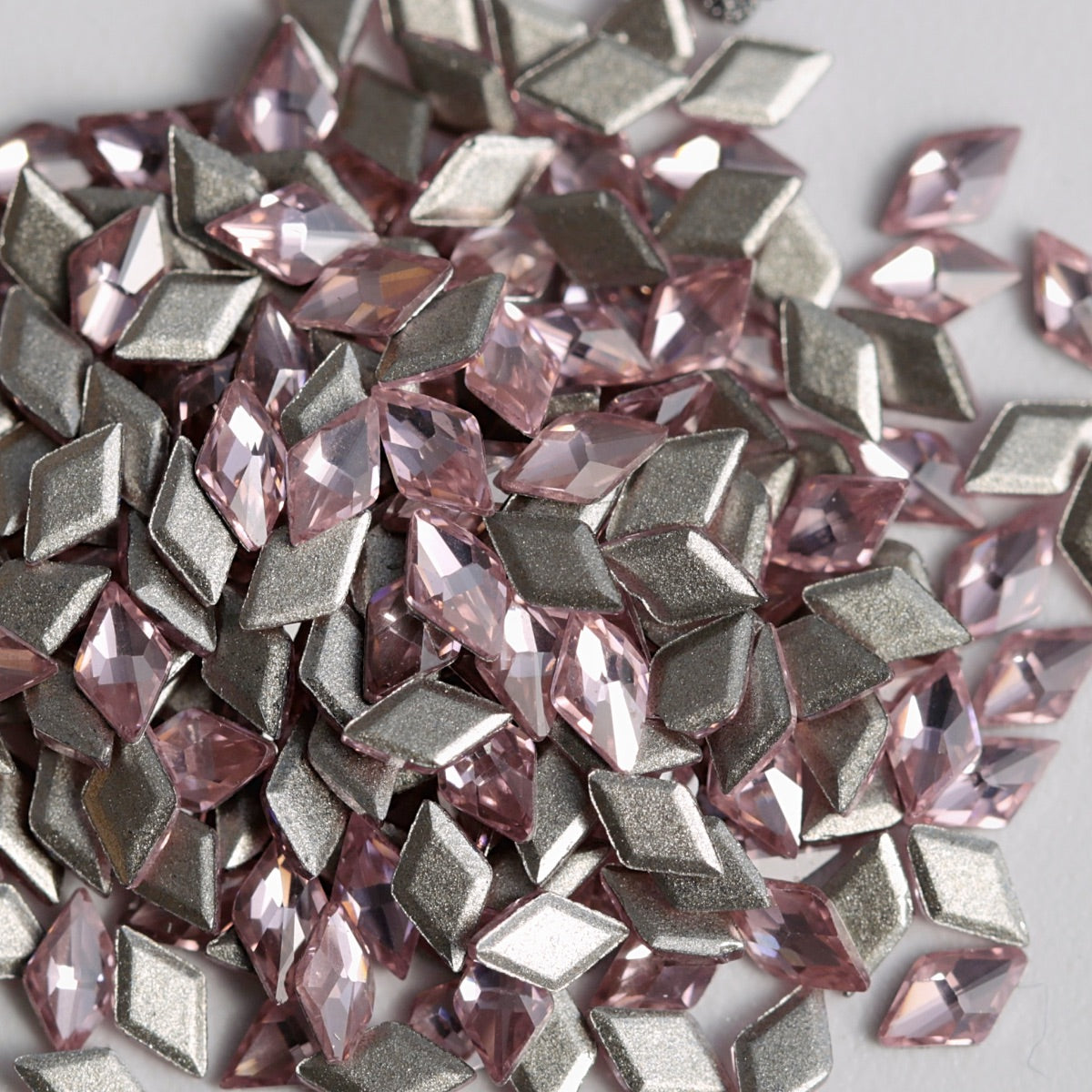 A detailed close-up of Light Rose rhombus-shaped zircon crystals arranged together with their reflective silver foil backings visible. The delicate pink color and sharp geometric facets add a sophisticated, multidimensional sparkle, making these crystals suitable for nail art, jewelry, accessories, and decorative craft projects.