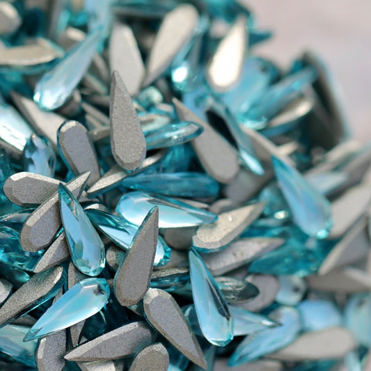 Pile of aquamarine glass flatback raindrop rhinestones with silver foil backing and faceted sparkle on light background for nail art