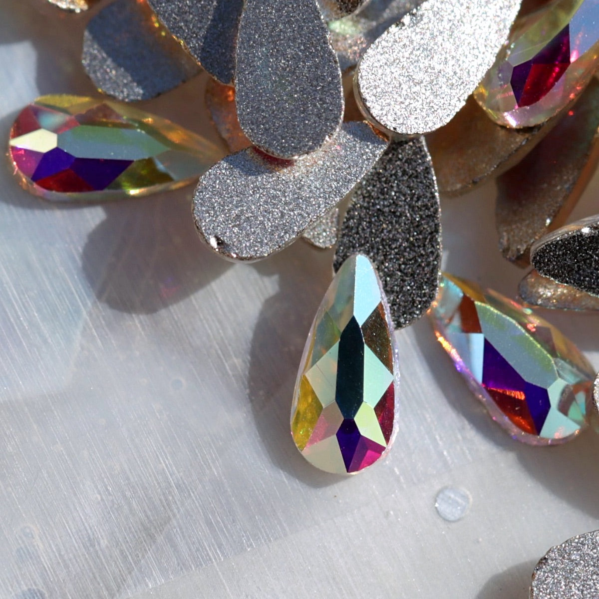 Crystal AB glass flatback teardrop crystals with a clear base and Aurora Borealis coating. Their rounded faceted droplet shape reflects blue, pink, purple, green, gold and silver flashes, making them ideal for crystal flowers, bridal nails, mermaid designs, winter manicures and elegant nail-art accents.
