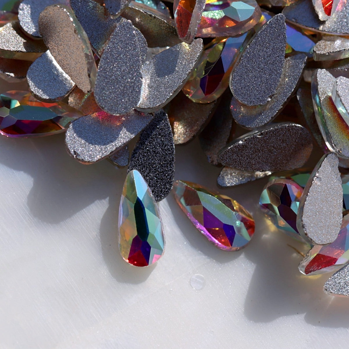 A close-up of iridescent rhinestone craft gems, including glittery silver teardrop shapes and faceted teardrop crystals with rainbow reflections, scattered on a light surface.