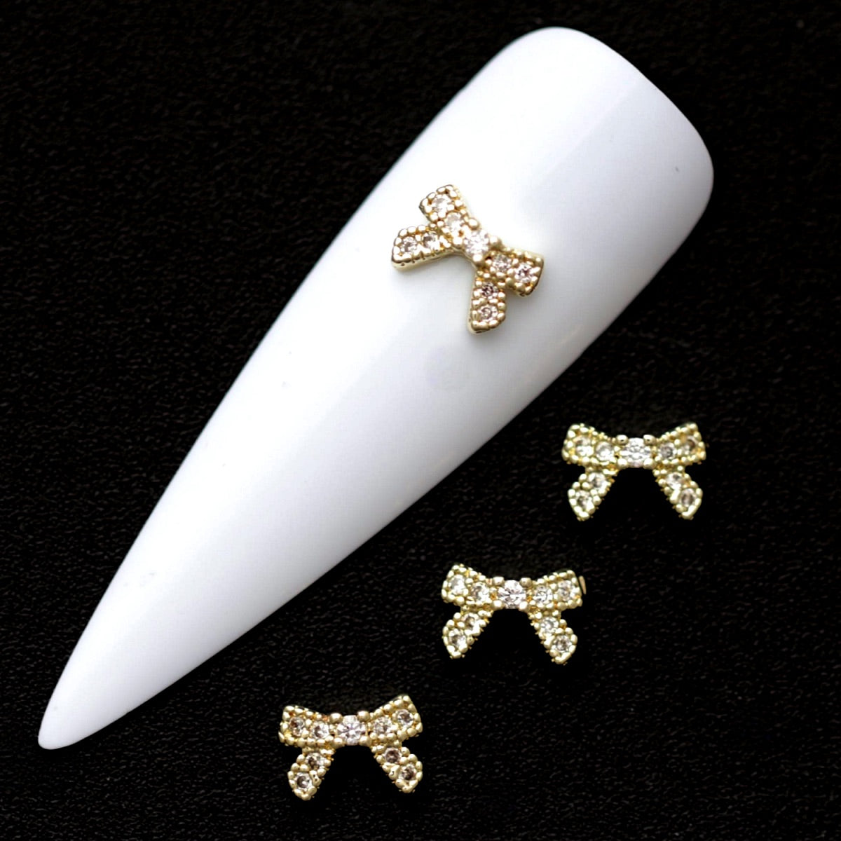 Gold 7×5 mm bow-shaped nail charms paved with crystal CZ stones displayed on a white nail tip against a black background