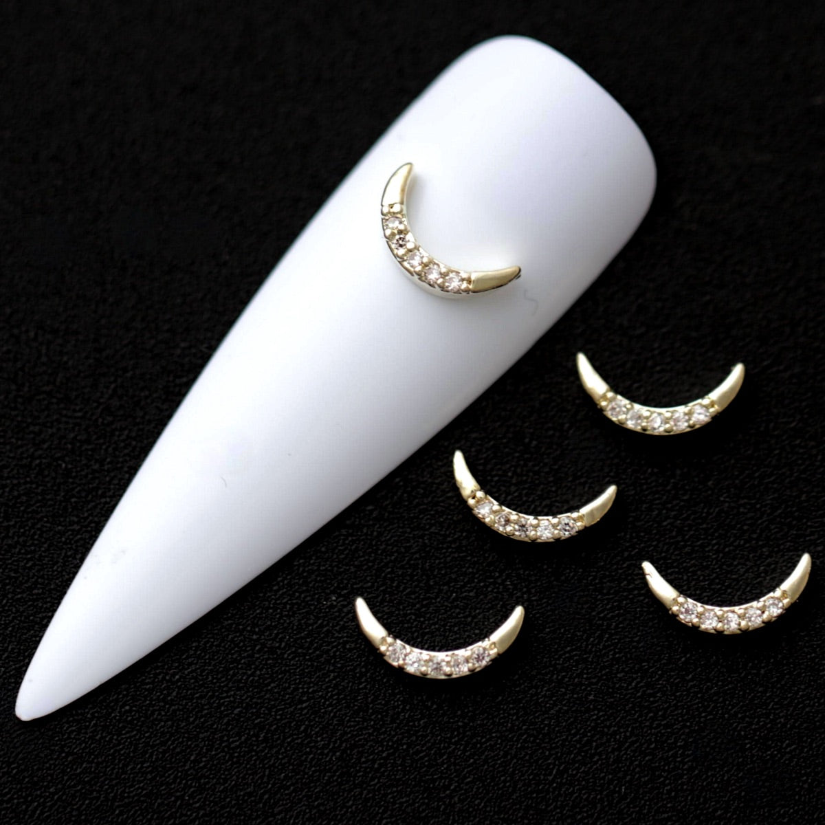 Gold crescent moon nail charms paved with crystal cubic zirconia on a white nail tip against a black background