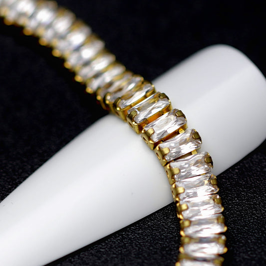 Close-up of gold cup chain with 4×2 mm clear zirconia baguette rhinestones displayed around a white nail tip on a black background