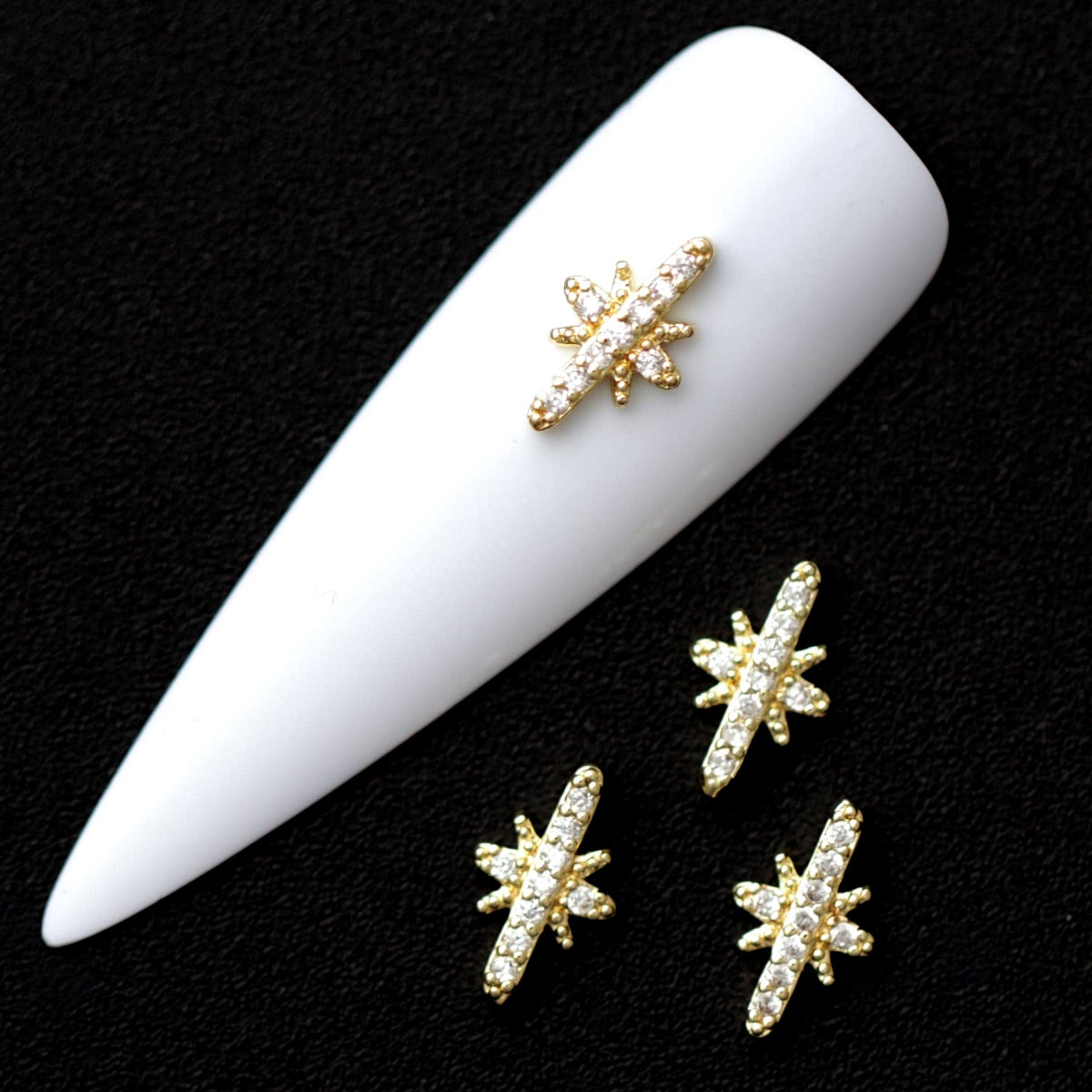 Gold 10×6 mm morning star nail charms paved with crystal CZ stones displayed on a white nail tip against a black background