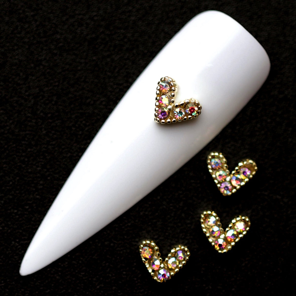 Gold 7×6 mm rope-style heart nail charms paved with iridescent CZ stones displayed on a white nail tip against a black background
