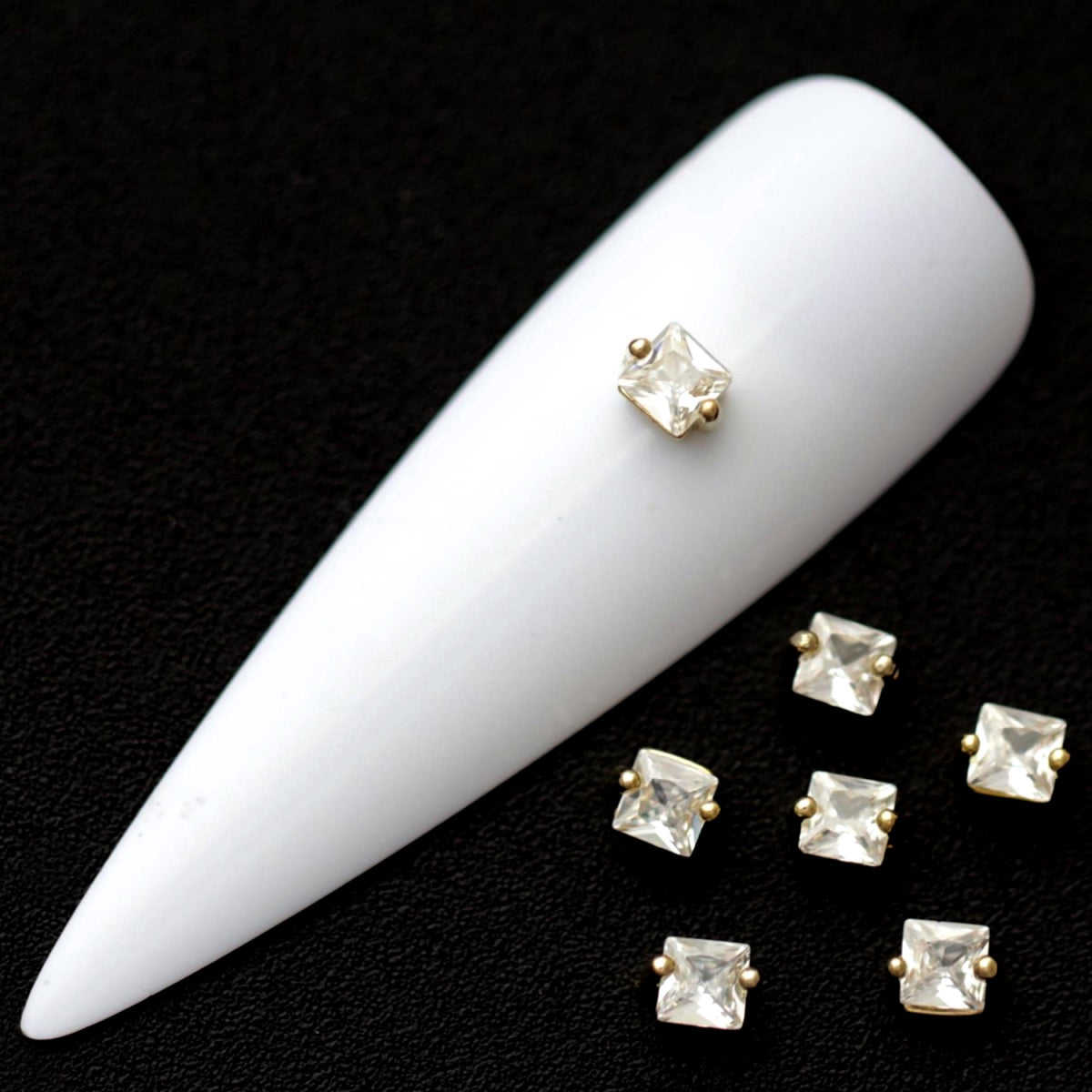 3 mm clear square cubic zirconia nail charms in gold settings displayed on a white nail tip against a black background