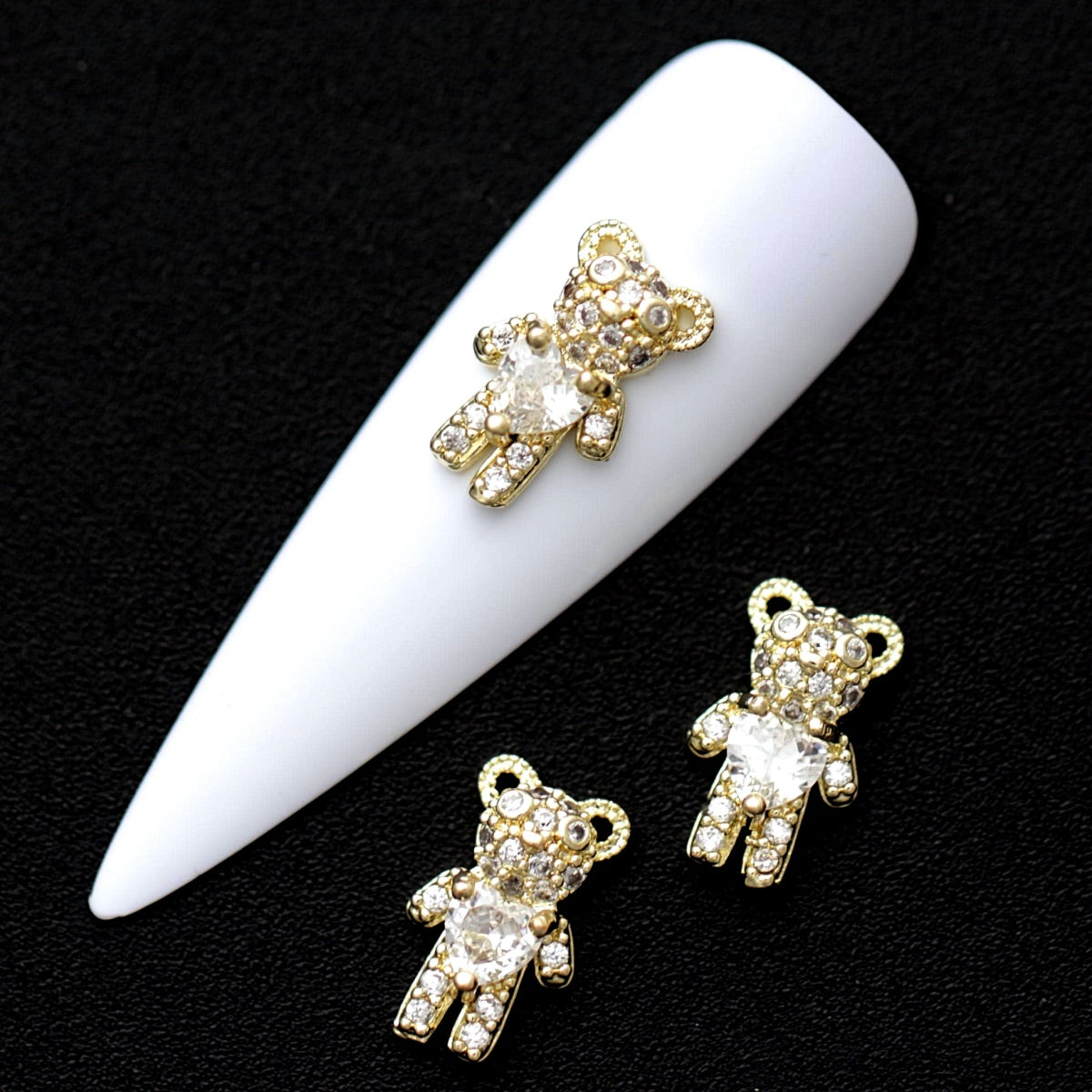 Gold 12×7 mm teddy bear nail charms with heart-shaped crystal CZ stones displayed on a white nail tip against a black background