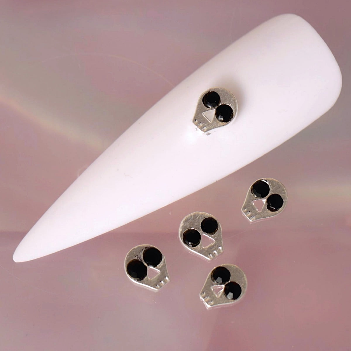 Halloween skull nail charm with a metallic silver-tone finish, glossy black eye details, a small triangular nose and subtle teeth markings. This compact spooky decoration is ideal for gothic manicures, horror-inspired designs, Halloween nail art and dark accent nails.