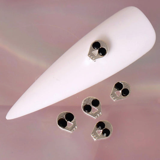 Halloween skull nail charm with a metallic silver-tone finish, glossy black eye details, a small triangular nose and subtle teeth markings. This compact spooky decoration is ideal for gothic manicures, horror-inspired designs, Halloween nail art and dark accent nails.